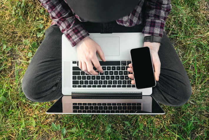 A man in a park on the grass sits and works at a laptop, holds a smartphone with his hand, top view.