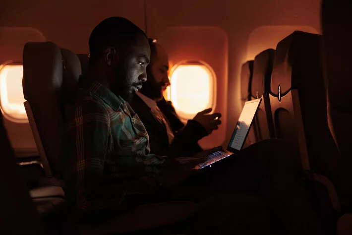 Freelancer passenger working on laptop during sunset, flying by airplane on commercial flight with international airline service. Male tourist travelling on work trip or holiday.