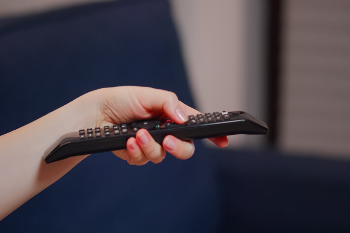 Close up of caucasian hand with TV remote switching channels while sitting on couch. Young person using technology finding movie to watch and relax after dinner, laying on sofa.